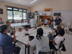 Em uma sala com uma grande janela, cerca de 10 pessoas em torno de mesas, cada uma segura um vaso de plantas e um pincel.